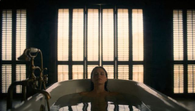 Backlit woman reclining in vintage freestanding bathtub with brass faucet and shutters
