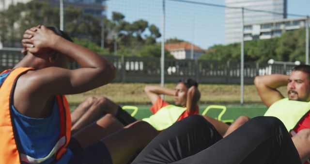 A group of athletes is performing sit-ups outdoors in a sports field with netted surroundings under a clear sky. They wear brightly colored vests suggesting a team exercise context. This image is suitable for illustrating fitness routines, team sports training, health and wellness articles, and fitness program promotions.