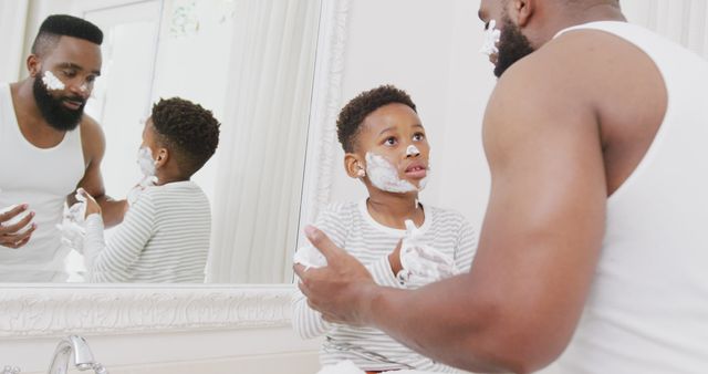 Father and young son engaging in shaving routine with foamy cream on faces. Ideal for content on parenting, family bonding activities, father-son relationships, early morning routines, and teaching new skills.