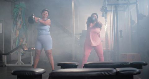 Diverse women swinging kettlebells in gritty gym with colorful mural and moody mist