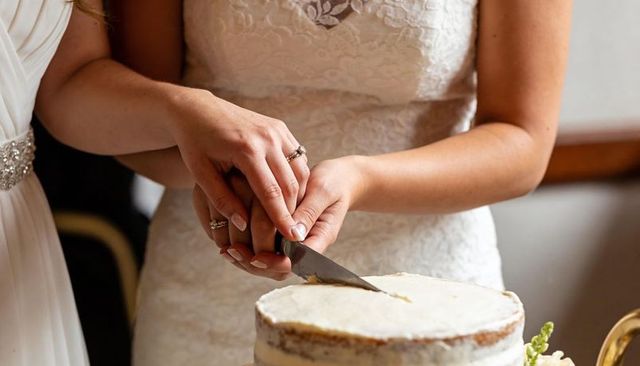 Two brides cutting wedding cake together wearing lace gowns and wedding rings