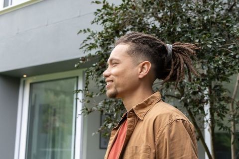 Young african american man standing in profile smiling with dreadlocks near modern home