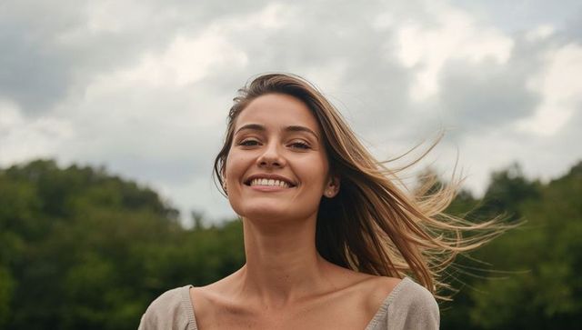 Smiling woman with windblown hair enjoying outdoors under overcast sky and treeline