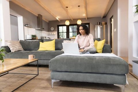 Non-binary person working on laptop in cozy open-plan living room with yellow cushions