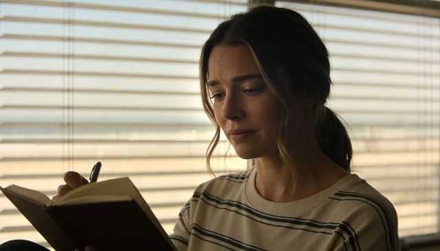 Contemplative woman writing in journal by window blinds with soft daylight, neutral tones