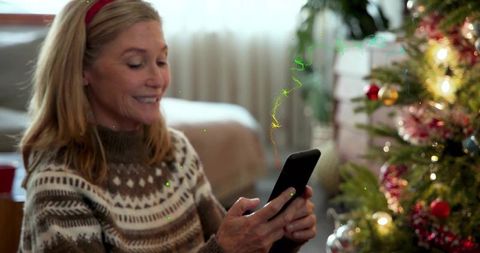 Smiling woman in knit sweater and red headband texting by decorated christmas tree