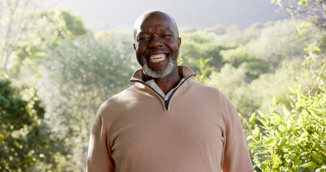Elderly man wears a casual pullover and beams a joyful smile while standing against a backdrop of dense greenery. This image can be used for promoting positive aging, outdoor activities for seniors, or emphasizing the benefits of a happy and healthy lifestyle in nature.