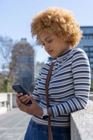 African american woman leaning on urban railing scrolling smartphone in striped top