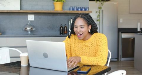 African american woman working remotely in modern kitchen wearing yellow sweater smiling