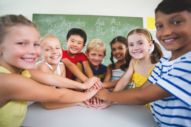This image shows a diverse group of school kids stacking hands in a classroom, symbolizing teamwork and unity. Ideal for educational materials, school websites, and articles on childhood development, teamwork, and diversity.