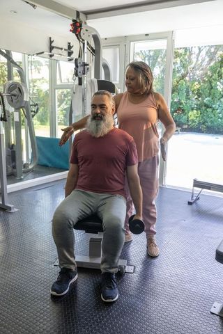 Senior man lifting dumbbell with african american woman spotting in home gym