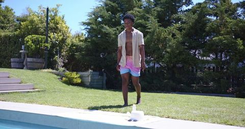 Sunlit backyard stroll shirtless man in open shirt colorful swim trunks heading to pool
