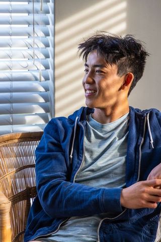 Teenage asian male sitting in wicker chair by sunlit window with striped blinds