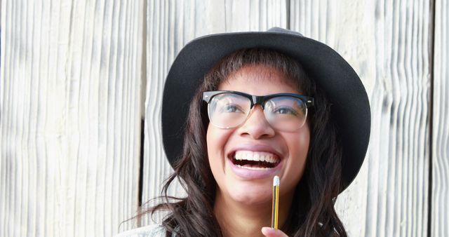 Afro-American woman smiling while holding a pencil and wearing a hat and glasses, standing against a light wooden background. Great for concepts of creativity, happiness, casual lifestyle, and outdoor activities. Ideal for use in advertisements, lifestyle blogs, and design thinking inspirations.