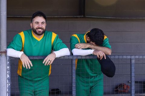Smiling and dejected baseball teammates leaning on dugout railing wearing green jerseys