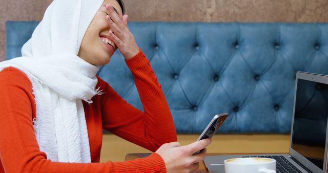 Muslim Woman Laughing While Holding Smartphone Near Laptop in Cafe - Download Free Stock Images Pikwizard.com