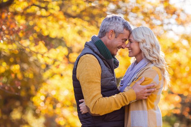 Romantic senior couple standing face to face, embracing in a park during autumn. The golden leaves and warm clothing suggest a cozy, intimate moment. Perfect for use in advertisements, greeting cards, or articles about love, relationships, and enjoying life in later years.