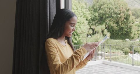 African American Woman Smiling While Reading Letter by Sunlit Sliding Glass Door