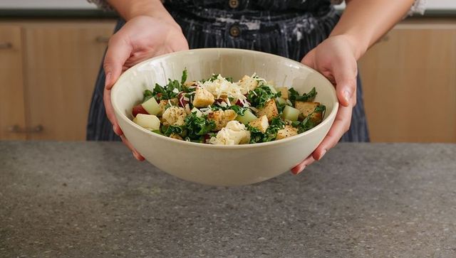 Home cook holding large ceramic bowl of fresh kale salad with croutons and shaved cheese