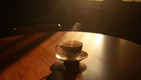 Steaming floral china teacup sitting on wooden table while sunlight casts warm shadows