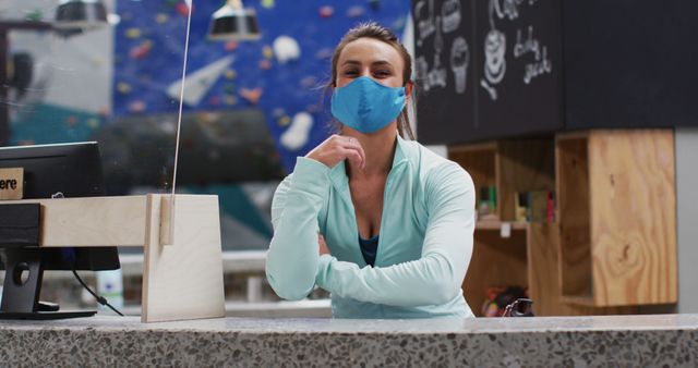 Fitness instructor standing at reception desk in climbing gym wearing protective mask. Useful for content about health and safety measures during fitness activities, gym precautions during pandemics, and fitness professionals. Enhances visuals in articles discussing the impact of COVID-19 on gym environments and climbing gyms.