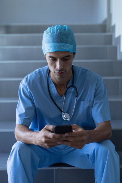 Male surgeon in blue scrubs and a surgical cap sitting on a hospital staircase, using a mobile phone. He has a stethoscope around his neck, indicating his medical profession. This image can be used to depict healthcare professionals taking a break, using technology for communication, or the daily life of medical staff in a hospital environment.