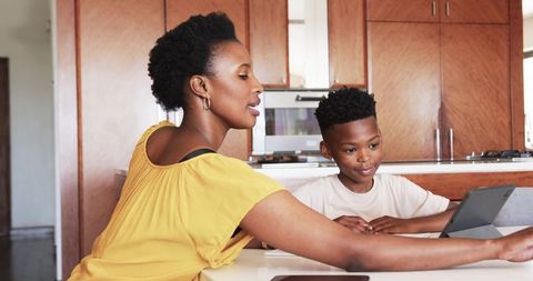 African American mother guiding son with tablet at sunlit kitchen table for learning