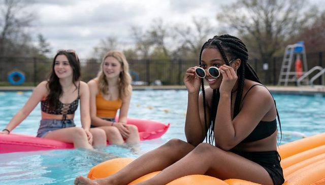 Young black woman adjusting sunglasses while floating on orange pool float with friends