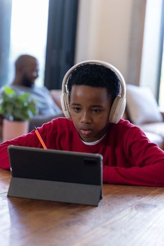 Young boy wearing headphones doing tablet homework at home with father in background