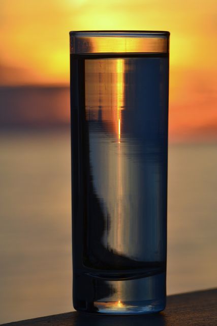 Tall glass filled with water reflecting a vibrant sunset sky colors. Useful for illustrating tranquility, peaceful evening moments, and the beauty of nature. Ideal for websites, blog posts, or advertisements focusing on relaxation, mindfulness, and well-being.