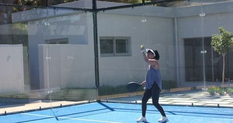 Young woman serving on outdoor padel court with glass walls and bright blue surface