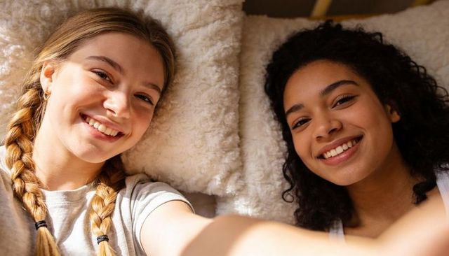Two young women smiling while taking cozy bedside selfie with textured cream pillows