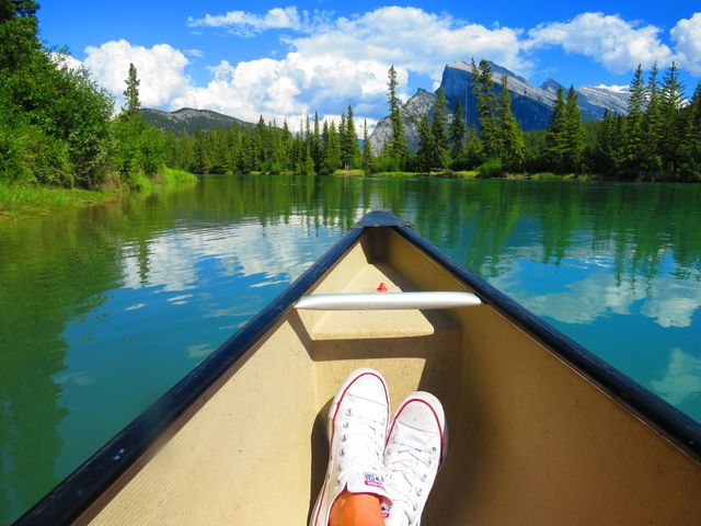 Perfect for promoting serene and relaxing outdoor adventures, this image shows the front view from inside a canoe on a calm lake surrounded by pine trees and mountains. The sky is bright blue with some clouds, making it ideal for advertising outdoor gear, travel brochures, leisure activities, and camping holidays.