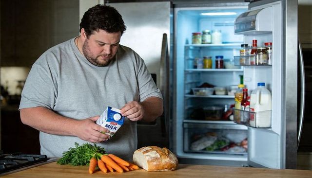 Man reading milk label at kitchen counter with open fridge, bread and fresh vegetables