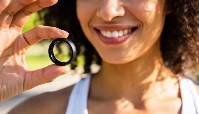 Smiling woman holding matte black ring closeup in sunlit park, modern accessory lifestyle