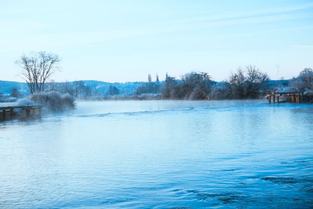 This image depicts a tranquil winter scene featuring a calm lake with a mist hovering over its surface. Distant trees partially covered in frost add depth and a serene feel to the landscape. The setting is ideal for projects related to nature, winter, tranquility, and outdoor activities. Use this image in environmental campaigns, travel brochures, outdoor adventure advertisements, or scenic backgrounds to evoke a sense of calm and peace.