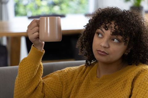 African american woman holding beige mug and looking up while relaxing in mustard sweater