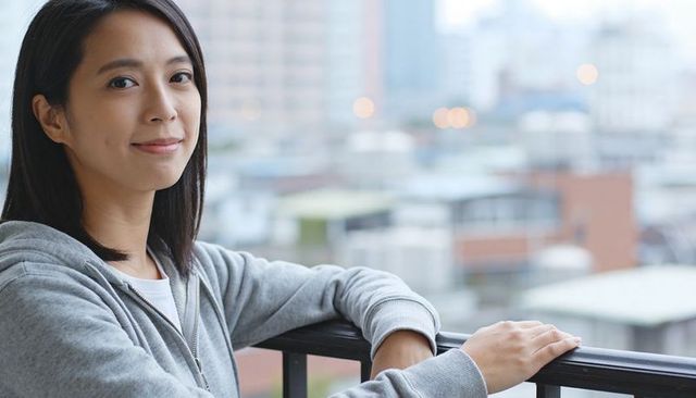 Asian woman leaning on balcony railing in grey hoodie with soft urban bokeh background