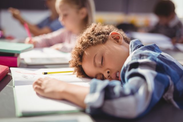 Young boy sleeping on his desk in a classroom, surrounded by books and pencils. Ideal for illustrating concepts of childhood fatigue, education challenges, and the importance of rest for students. Suitable for educational materials, parenting blogs, and articles on student well-being.