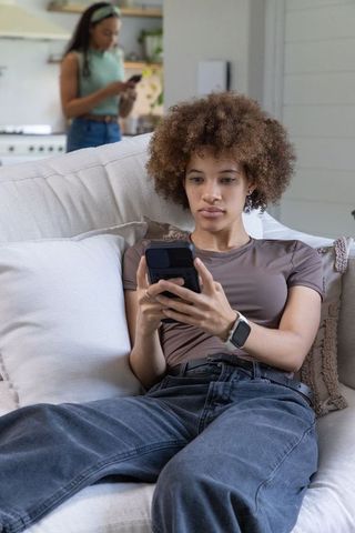 Young woman reclining on sofa checking smartphone while roommate using phone in background