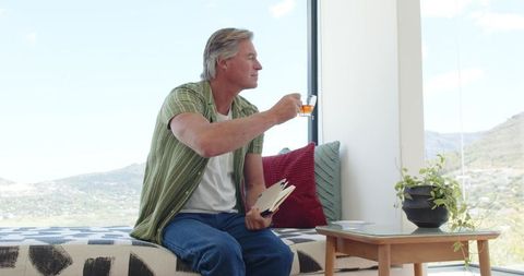Mature man sipping tea and reading on cozy window daybed with mountain view