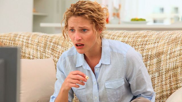 Female with curly hair sitting on a couch in a cozy living room while watching a scary movie. Her facial expression reflects fear, adding drama to the scene. Suitable for use in articles or images related to home entertainment, movie reactions, living room decor, or emotional experiences.