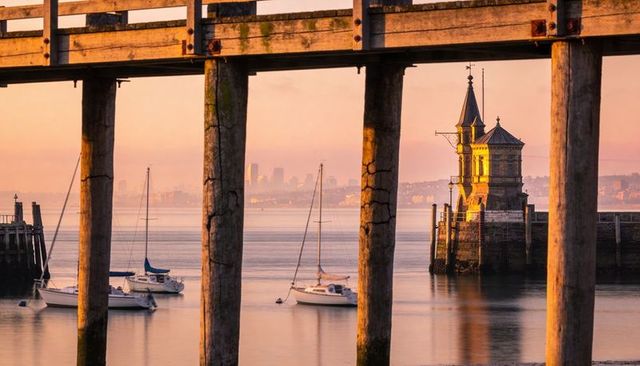 Golden-hour harbor framed by weathered pier posts with moored sailboats and lighthouse tower