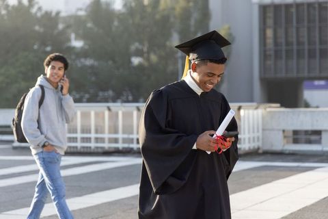 African american graduate checking smartphone while holding diploma on campus plaza