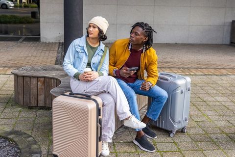Young diverse travelers waiting with suitcases on urban bench holding coffee and phone