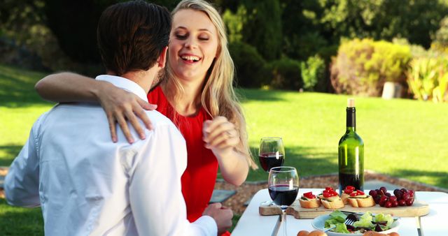 Happy couple enjoying romantic picnic in park. Woman in red dress is hugging her partner while sitting at table with wine and appetizers. Ideal for promoting romance, outdoor activities, or fine wine.