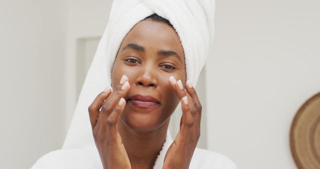 Image of happy african american woman in robe moisturizing face. beauty treatment and skin care routine concept.