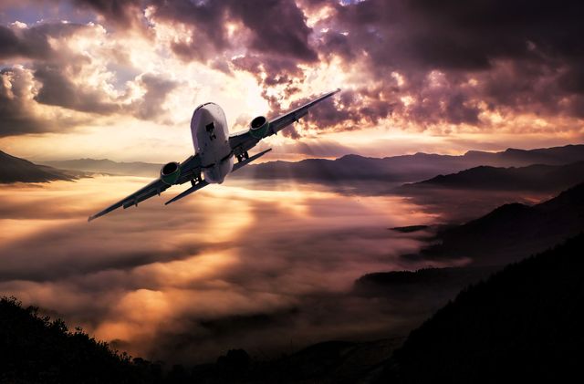 Depicts a commercial aircraft soaring above a dense layer of clouds with a dramatic sunset in the background and mountainous terrain below. Ideal for travel promotions, aviation industry advertisements, and inspirational positioning related to flight. Scenic imagery can be used in brochures, banners, and websites about air journeys or scenic destinations.