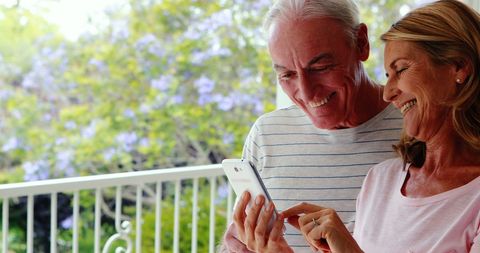 Joyful Elderly Couple Browsing Smartphone on Sunlit Balcony