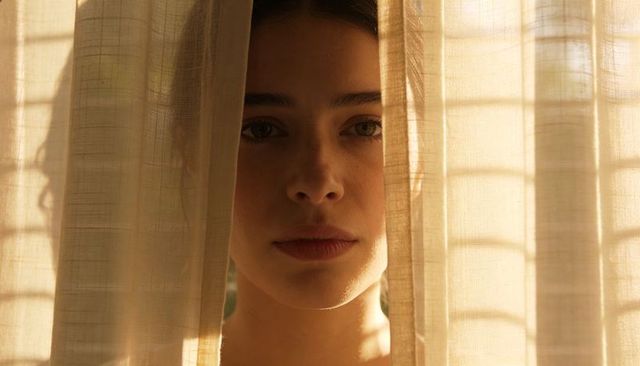 Young woman gazing through sunlit sheer curtains, closeup portrait with grid shadows
